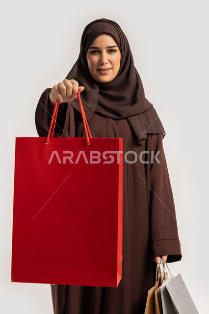 تعابير وايماءات السرور والسعادة بتقديم الهدية، الاستفادة من العروض والخصومات، إمرأة سعودية تقوم بالتسوق وشراء المستلزمات, بورتريه لسيدة عربية خليجية اماراتية مبتمسة ترتدي الحجاب والعباءة البنية تحمل بيدها أكياس كرتون ملونه، خلفية بيضاء