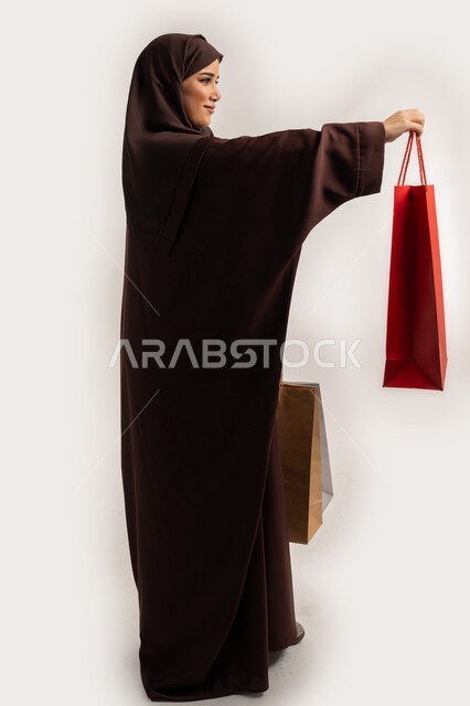 تقديم الهدية بايماءات السرور والسعادة، الاستفادة من العروض والخصومات, إمرأة سعودية تقوم بالتسوق وشراء المستلزمات, بورتريه من الجانب لسيدة عربية خليجية اماراتية مبتمسة ترتدي الحجاب والعباءة البنية تحمل بيدها أكياس كرتون ملونه، الجسم بالطول الكامل، خلفية بيضاء