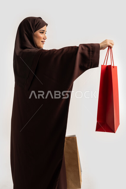 الاستفادة من العروض والخصومات, تقديم الهدية بايماءات وتعابير السرور والسعادة، إمرأة سعودية تقوم بالتسوق وشراء المستلزمات, بورتريه من الجانب لسيدة عربية خليجية اماراتية مبتمسة ترتدي الحجاب والعباءة البنية تحمل بيدها أكياس كرتون ملونه، خلفية بيضاء