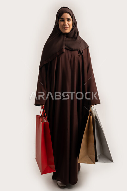 الوقوف باستقامة بايماءات السرور والسعادة، الاستفادة من العروض والخصومات, إمرأة سعودية تقوم بالتسوق وشراء المستلزمات والهدايا, بورتريه لسيدة عربية خليجية اماراتية مبتمسة ترتدي الحجاب والعباءة البنية تحمل بيدها أكياس كرتون ملونه، الجسم بالطول الكامل، خلفية بيضاء