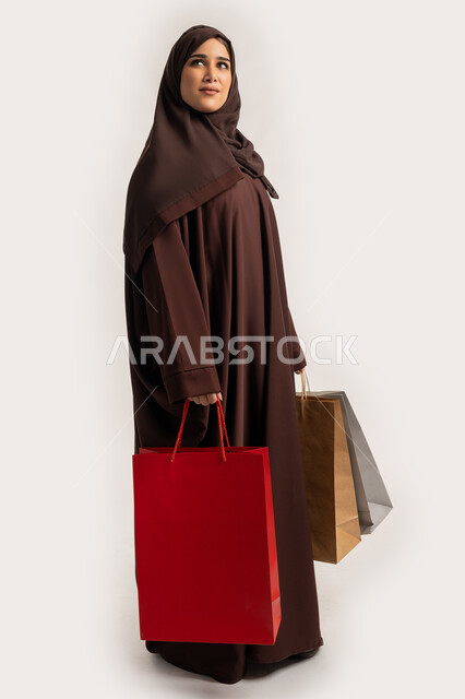 النظرالى الاعلى بايماءات السرور والسعادة، الاستفادة من العروض والخصومات, إمرأة سعودية تقوم بالتسوق وشراء المستلزمات والهدايا, بورتريه لسيدة عربية خليجية اماراتية مبتمسة ترتدي الحجاب والعباءة البنية تحمل بيدها أكياس كرتون ملونه، الجسم بالطول الكامل، خلفية بيضاء