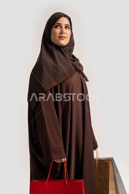 النظرالى الاعلى بايماءات وتعابير السرور والسعادة، الاستفادة من العروض والخصومات, إمرأة سعودية تقوم بالتسوق وشراء المستلزمات والهدايا, بورتريه لسيدة عربية خليجية اماراتية مبتمسة ترتدي الحجاب والعباءة البنية تحمل بيدها أكياس كرتون ملونه، خلفية بيضاء
