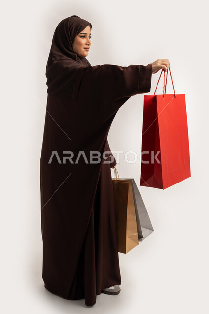 تقديم الهدية بايماءات السرور والسعادة، الاستفادة من العروض والخصومات, إمرأة سعودية تقوم بالتسوق وشراء المستلزمات, بورتريه من الجانب لسيدة عربية خليجية اماراتية مبتمسة ترتدي الحجاب والعباءة البنية تحمل بيدها أكياس كرتون ملونه، الجسم بالطول الكامل، خلفية بيضاء