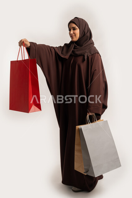 Expressions of pleasure and happiness in presenting a gift, taking advantage of offers and discounts, a Saudi woman shopping and buying supplies, side portrait of a smiling Emirati Gulf Arab woman wearing a hijab and a brown abaya holding colorful cardboard bags, full body, white background