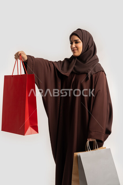 تعابير وايماءات السرور والسعادة بتقديم الهدية، الاستفادة من العروض والخصومات، إمرأة سعودية تقوم بالتسوق وشراء المستلزمات, بورتريه من الجانب لسيدة عربية خليجية اماراتية مبتمسة ترتدي الحجاب والعباءة البنية تحمل بيدها أكياس كرتون ملونه، خلفية بيضاء