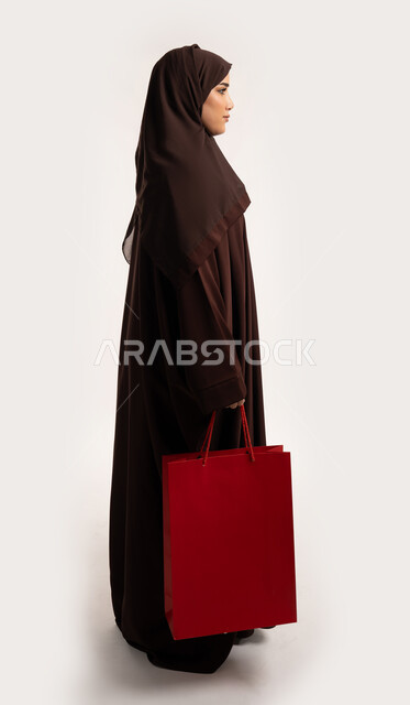 ايماءات المشي والذهاب لمكان ما، الاستفادة من العروض والخصومات، إمرأة سعودية تقوم بالتسوق وشراء المستلزمات, بورتريه من الجانب لسيدة عربية خليجية اماراتية مبتمسة ترتدي الحجاب والعباءة البنية تحمل بيدها أكياس كرتون ملونه، الجسم بالطول الكامل، خلفية بيضاء