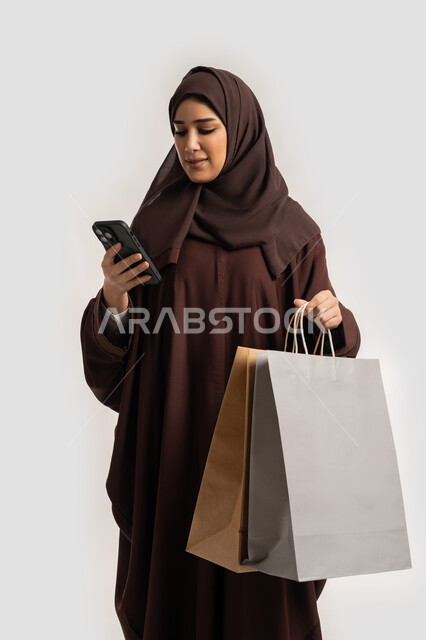 تعابير السرور والسعادة، استخدام التكنولوجيا في الحياة اليومية، إمرأة سعودية تقوم بالتسوق وشراء المستلزمات والهدايا عبر الانترنت، بورتريه لسيدة عربية خليجية اماراتية ترتدي الحجاب والعباءة تمسك بيدها الهاتف المحمول وأكياس التسوق، خلفية بيضاء