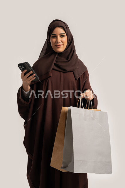 النظر الى الكاميرا بتعابير السرور والسعادة، استخدام التكنولوجيا في الحياة اليومية، إمرأة سعودية تقوم بالتسوق وشراء المستلزمات والهدايا عبر الانترنت، بورتريه لسيدة عربية خليجية اماراتية ترتدي الحجاب والعباءة تمسك بيدها الهاتف المحمول وأكياس التسوق، خلفية بيضاء