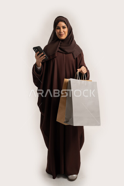 النظر الى الكاميرا بتعابير السعادة، استخدام التكنولوجيا في الحياة اليومية، إمرأة سعودية تقوم بالتسوق وشراء المستلزمات والهدايا عبر الانترنت، بورتريه لسيدة عربية خليجية اماراتية ترتدي الحجاب والعباءة تمسك الهاتف المحمول وأكياس التسوق، الجسم بالطول الكامل، خلفية بيضاء