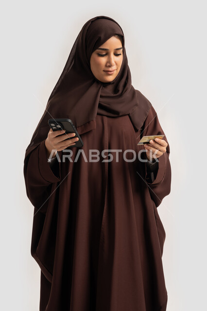 Possibility of purchasing and paying through mobile programs and applications, portrait of a smiling Emirati Gulf Arab woman wearing hijab and abaya holding a mobile phone and a bank credit card, a Saudi woman making payments online, using modern technical devices, white background