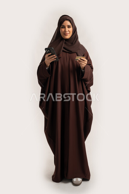 Using modern technical devices, the ability to purchase and pay through mobile programs and applications, a portrait of an Arab Gulf Emirati woman wearing a hijab and an abaya, holding a mobile phone and a bank credit card in her hand, a Saudi woman making payments online, full body, white background