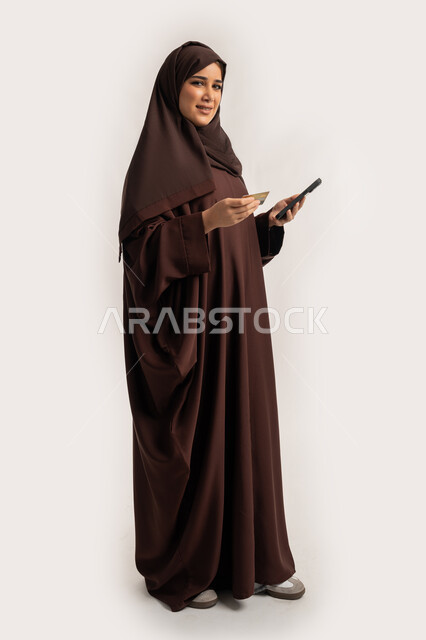 Using modern technical devices, the ability to purchase and pay through mobile programs and applications, a portrait of an Arab Gulf Emirati woman wearing a hijab and an abaya, holding a mobile phone and a bank credit card in her hand, a Saudi woman making payments online, full body, white background