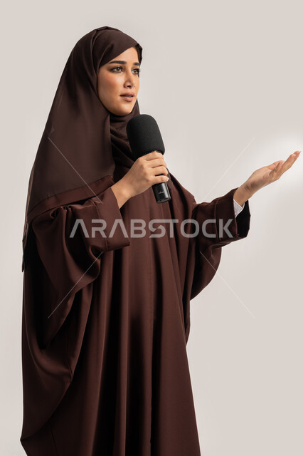 مهارات التحدث والحوار، فن الالقاء والخطابة، إمرأة سعودية تقوم بتقديم عرض فني غنائي وفقرات ترفيهية باستخدام مكبر الصوت، بورتريه لشابة عربية خليجية اماراتية محجبة ترتدي العباءة البنية تمسك بيدها المايكروفون بإيماءات الاندماج والتركيز، خلفيه بيضاء