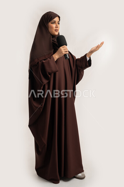 مهارات التحدث والحوار، فن الالقاء والخطابة، إمرأة سعودية تقوم بتقديم عرض فني غنائي وفقرات ترفيهية باستخدام مكبر الصوت، بورتريه لشابة عربية خليجية اماراتية محجبة ترتدي العباءة البنية تمسك المايكروفون بإيماءات الاندماج والتركيز، الجسم بالطول الكامل، خلفيه بيضاء