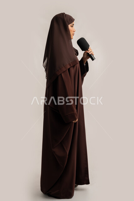مهارات التحدث والحوار، فن الالقاء والخطابة، إمرأة سعودية تقوم بتقديم عرض فني غنائي وفقرات ترفيهية باستخدام مكبر الصوت، بورتريه من الجانب لشابة عربية خليجية اماراتية محجبة ترتدي العباءة تمسك المايكروفون بإيماءات الاندماج والتركيز، الجسم بالطول الكامل، خلفيه بيضاء
