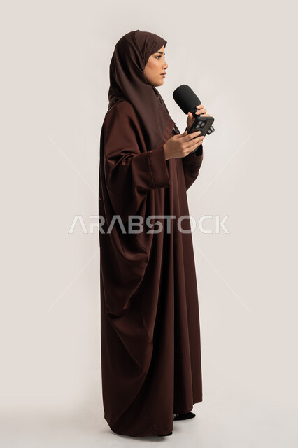 التواصل الفعال وتبادل الحوار والنقاش، إقامة ورش العمل والدورات التدريبية، إمرأة سعودية تقوم بالقراءة من الهاتف المحمول، فن الالقاء والخطابة، بورتريه لشابة عربية خليجية اماراتية محجبة ترتدي العباءة تمسك مكبر الصوت تلقي الخطابات، الجسم بالطول الكامل، خلفية بيضاء