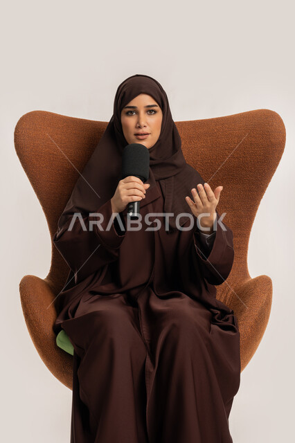 إيماءات الاندماج والتركيز، مهارة التحدث والحوار، إمرأة سعودية تجلس على الكرسي تقوم بتقديم ورش العمل والدورات التدريبية باستخدام مكبر الصوت، بورتريه من الجانب لشابة عربية خليجية اماراتية محجبة ترتدي العباءة تمسك المايكروفون تتقن فن الالقاء والخطابة، خلفيه بيضاء