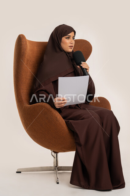 تقديم ورش العمل والدورات التدريبية، فن التحدث والخطابة، إمرأة سعودية تقوم بالقاء الخطابات من الورقة بإيماءات الثقة بالنفس، التواصل الفعال وتبادل الحوار والنقاش، بورتريه لشابة عربية خليجية اماراتية محجبة ترتدي العباءة تجلس على الكرسي تمسك مكبر الصوت، خلفية بيضاء
