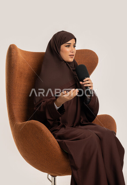 Mastering the art of public speaking and oratory, speaking and dialogue skills, a Saudi woman sitting on a chair presenting workshops and training courses using a loudspeaker, a side portrait of a veiled young Arab Gulf Emirati woman wearing an abaya holding the microphone with gestures of integration and focus, white background
