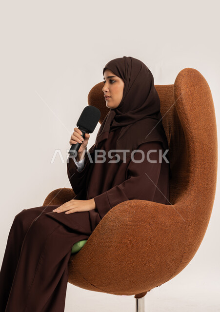 مهارة التحدث والحوار، اتقان فن الالقاء والخطابة، إمرأة سعودية تجلس على الكرسي تقوم بتقديم ورش العمل والدورات التدريبية باستخدام مكبر الصوت، بورتريه من الجانب لشابة عربية خليجية اماراتية محجبة ترتدي العباءة تمسك المايكروفون بإيماءات الاندماج والتركيز، خلفيه بيضاء