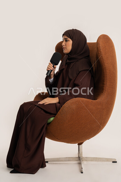 مهارة التحدث والحوار، اتقان فن الالقاء والخطابة، إمرأة سعودية تجلس على الكرسي تقوم بتقديم ورش العمل والدورات التدريبية باستخدام مكبر الصوت، بورتريه من الجانب لشابة عربية خليجية اماراتية محجبة ترتدي العباءة تمسك المايكروفون بإيماءات الاندماج والتركيز، خلفيه بيضاء