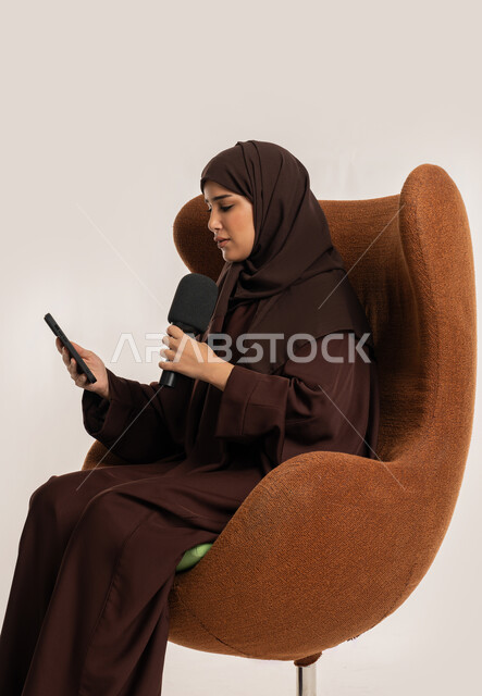 التواصل الفعال وتبادل الحوار والنقاش، إقامة ورش العمل والدورات التدريبية، إمرأة سعودية تجلس على الكرسي تقوم بالقراءة من الهاتف المحمول، فن الالقاء والخطابة، بورتريه من الجانب لشابة عربية خليجية اماراتية محجبة ترتدي العباءة تمسك مكبر الصوت تلقي الخطابات، خلفية بيضاء