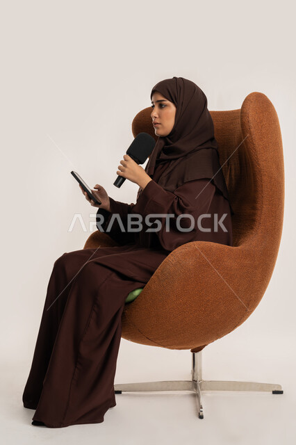 التواصل الفعال وتبادل الحوار والنقاش، إقامة ورش العمل والدورات التدريبية، إمرأة سعودية تجلس على الكرسي تقوم بالقراءة من الهاتف المحمول، فن الالقاء والخطابة، بورتريه من الجانب لشابة عربية خليجية اماراتية محجبة ترتدي العباءة تمسك مكبر الصوت تلقي الخطابات، خلفية بيضاء
