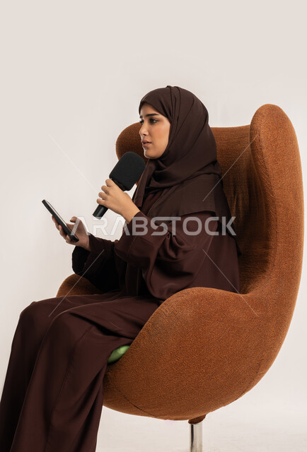 تبادل الحوار والنقاش والتواصل الفعال، إقامة ورش العمل والدورات التدريبية، إمرأة سعودية تجلس على الكرسي تقوم بالقراءة من الهاتف المحمول، فن الالقاء والخطابة، بورتريه من الجانب لشابة عربية خليجية اماراتية محجبة ترتدي العباءة تمسك مكبر الصوت تلقي الخطابات، خلفية بيضاء