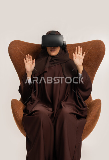 الاندماج والتفاعل ومحاكاة العالم الخيالي، بورتريه لإمرأة عربية خليجية اماراتية ترتدي الحجاب والعباءة ونظارات الواقع الافتراضي نجلس على الكرسي تقوم بمد يديها الى الامام بتعابير صد شيء ما والدفاع عن النفس، شابة سعودية تستخدم نظارة الفي آر، خلفية بيضاء