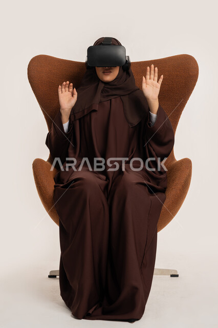 الاندماج والتفاعل ومحاكاة العالم الخيالي، بورتريه لإمرأة عربية خليجية اماراتية ترتدي الحجاب والعباءة ونظارات الواقع الافتراضي نجلس على الكرسي تقوم بمد يديها الى الامام بتعابير صد شيء ما والدفاع عن النفس، شابة سعودية تستخدم نظارة الفي آر، خلفية بيضاء
