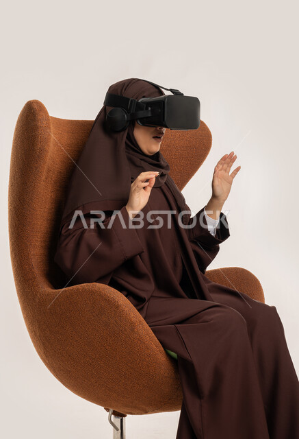 محاكاة العالم الخيالي والاندماج والتفاعل، بورتريه من الجانب لإمرأة عربية خليجية اماراتية ترتدي الحجاب والعباءة ونظارات الواقع الافتراضي نجلس على الكرسي تقوم بمد يديها الى الامام بتعابير صد شيء ما والدفاع عن النفس، شابة سعودية تستخدم نظارة الفي آر، خلفية بيضاء