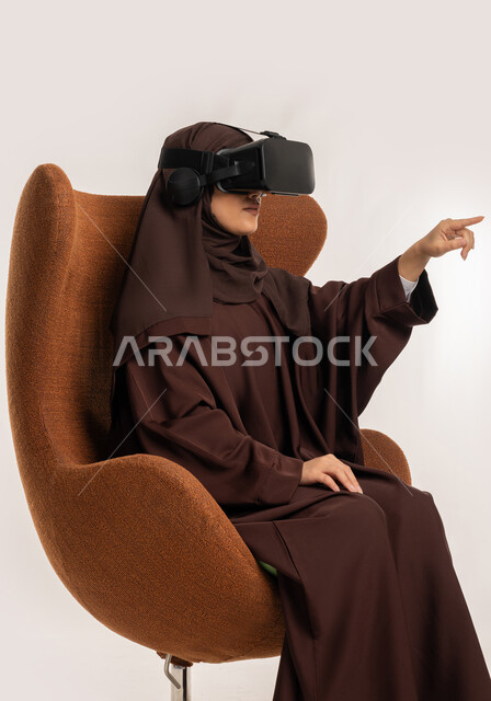 محاولة لمس شيء ما، تعابير الاندماج والتفاعل، بورتريه من الجانب لإمرأة عربية خليجية اماراتية ترتدي الحجاب والعباءة ونظارات الواقع الافتراضي تجلس على الكرسي تقوم بمد يديها الى الامام ومحاكاة العالم الخيالي، شابة سعودية تستخدم نظارة الفي آر، خلفية بيضاء