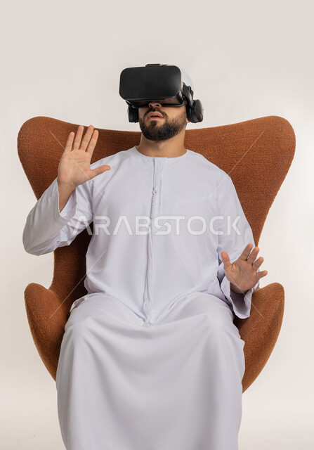 محاكاة العالم الخيالي والاندماج والتفاعل، بورتريه لرجل عربي خليجي اماراتي يرتدي الكندورة والقبعة ونظارات الواقع الافتراضي يجلس على الكرسي يقوم بمد يديه الى الامام بتعابير صد شيء ما والدفاع عن النفس، استخدام التقنية الحديثة المتطورة والتكنولوجيا، خلفية بيضاء