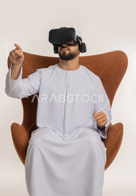 استخدام التقنية الحديثة المتطورة والتكنولوجيا، تعابير الاندماج والنقر والتفاعل، محاولة لمس شيء ما، بورتريه لرجل عربي خليجي اماراتي يرتدي القبعة والكندورة ونظارات الواقع الافتراضي يجلس على الكرسي يقوم برفع يديه ومحاكاة العالم الخيالي، خلفية بيضاء