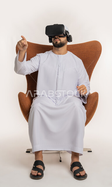 Using modern advanced technology and technology, expressions of integration, clicking and interaction, trying to touch something, portrait of an Arab Gulf Emirati man wearing a ghutra, kandura and virtual reality glasses sitting on a chair raising his hands and simulating the imaginary world, white background
