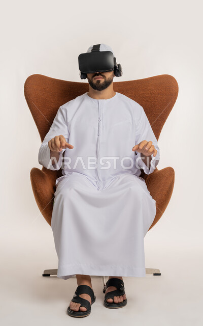 محاولة لمس شيء ما، استخدام التقنية الحديثة المتطورة والتكنولوجيا، تعابير الاندماج والنقر والتفاعل، بورتريه لرجل عربي خليجي اماراتي يرتدي القبعة والكندورة ونظارات الواقع الافتراضي يجلس على الكرسي يقوم برفع يديه ومحاكاة العالم الخيالي، خلفية بيضاء