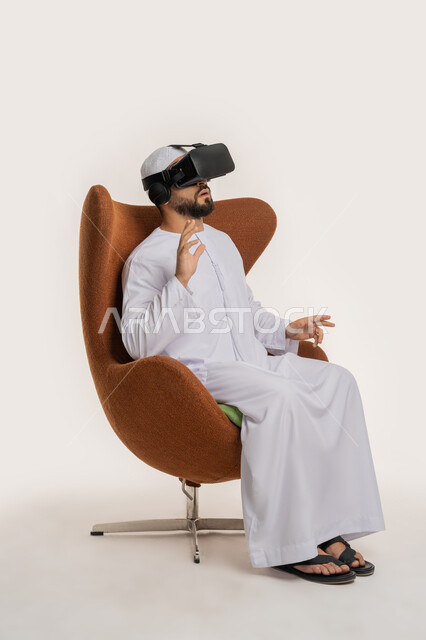 محاكاة العالم الخيالي والاندماج، بورتريه من الجانب لرجل عربي خليجي اماراتي يرتدي الكندورة والقبعة ونظارات الواقع الافتراضي يجلس على الكرسي يقوم بمد يديه الى الامام بتعابير صد شيء ما والدفاع عن النفس، استخدام التقنية الحديثة المتطورة والتكنولوجيا، خلفية بيضاء