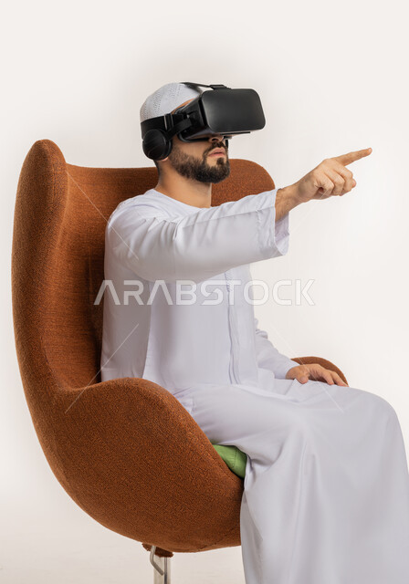 الاندماج والنقر والتفاعل، محاولة لمس شيء ما، بورتريه من الجانب لرجل عربي خليجي اماراتي يرتدي القبعة والكندورة ونظارات الواقع الافتراضي يجلس على الكرسي يقوم برفع يديه ومحاكاة العالم الخيالي، استخدام التقنية الحديثة المتطورة والتكنولوجيا، خلفية بيضاء