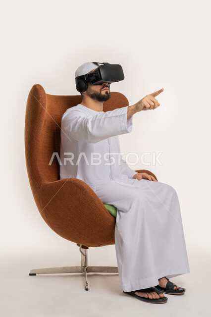 النقر والاندماج والتفاعل، محاولة لمس شيء ما، بورتريه من الجانب لرجل عربي خليجي اماراتي يرتدي القبعة والكندورة ونظارات الواقع الافتراضي يجلس على الكرسي يقوم برفع يديه ومحاكاة العالم الخيالي، استخدام التقنية الحديثة المتطورة والتكنولوجيا، خلفية بيضاء