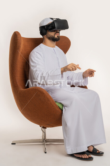 النقر والاندماج والتفاعل، محاولة لمس شيء ما، بورتريه من الجانب لرجل عربي خليجي اماراتي يرتدي القبعة والكندورة ونظارات الواقع الافتراضي يجلس على الكرسي يقوم برفع يديه ومحاكاة العالم الخيالي، استخدام التقنية الحديثة المتطورة والتكنولوجيا، خلفية بيضاء