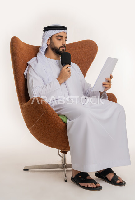 القاء الخطابات من الورقة بإيماءات الثقة بالنفس، التواصل الفعال وتبادل الحوار والنقاش، تقديم ورش العمل والدورات التدريبية، فن التحدث والخطابة، بورتريه لشاب عربي خليجي اماراتي يرتدي الكندورة والغترة يجلس على الكرسي يمسك مكبر الصوت، خلفية بيضاء