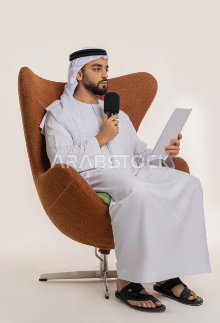 القاء الخطابات من الورقة بإيماءات الثقة بالنفس، التواصل الفعال وتبادل الحوار والنقاش، تقديم ورش العمل والدورات التدريبية، فن التحدث والخطابة، بورتريه لشاب عربي خليجي اماراتي يرتدي الكندورة والغترة يجلس على الكرسي يمسك مكبر الصوت، خلفية بيضاء
