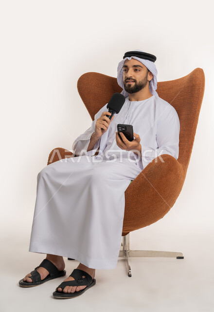 إلقاء الخطاب بإيماءات الثقة بالنفس، إقامة ورش العمل والدورات التدريبية، فن التحدث والخطابة، التواصل الفعال والنقاش، بورتريه لشاب عربي خليجي اماراتي يرتدي الكندورة والغترة يمسك مكبر الصوت يجلس على الكرسي تقوم بالقراءة من الهاتف المحمول، خلفية بيضاء