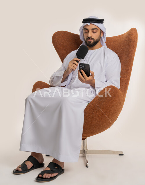 إقامة ورش العمل والدورات التدريبية، فن التحدث والخطابة، التواصل الفعال والنقاش، بورتريه لشاب عربي خليجي اماراتي يرتدي الكندورة والغترة يمسك مكبر الصوت يجلس على الكرسي تقوم بالقراءة من الهاتف المحمول، إلقاء الخطاب بإيماءات الثقة بالنفس، خلفية بيضاء