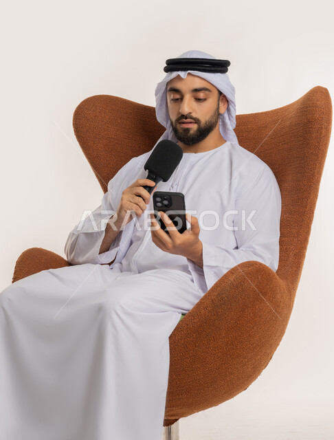 إلقاء الخطاب بإيماءات الثقة بالنفس، إقامة ورش العمل والدورات التدريبية، فن التحدث والخطابة، التواصل الفعال والنقاش، بورتريه لشاب عربي خليجي اماراتي يرتدي الكندورة والغترة يمسك مكبر الصوت يجلس على الكرسي تقوم بالقراءة من الهاتف المحمول، خلفية بيضاء