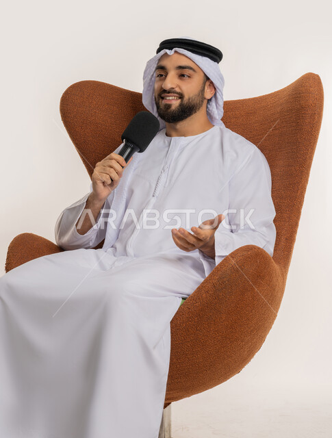 ايماءات وتعابير الاندماج والتركيز، اتقان فن الالقاء والخطابة، مهارات التحدث والحوار، بورتريه لشاب عربي خليجي اماراتي يرتدي الكندورة والغترة يجلس على الكرسي يقوم بتقديم ورش العمل والدورات التدريبية باستخدام مكبر الصوت، خلفيه بيضاء