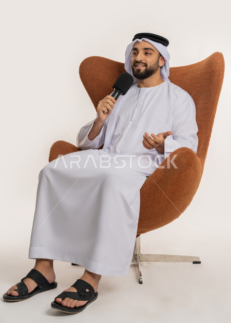 اتقان فن الالقاء والخطابة، مهارات التحدث والحوار، بورتريه لشاب عربي خليجي اماراتي يرتدي الكندورة والغترة يجلس على الكرسي يقوم بتقديم ورش العمل والدورات التدريبية باستخدام مكبر الصوت، ايماءات وتعابير الاندماج والتركيز، خلفيه بيضاء