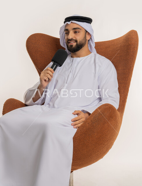ايماءات وتعابير الاندماج والتركيز، اتقان فن الالقاء والخطابة، مهارات التحدث والحوار، بورتريه لشاب عربي خليجي اماراتي يرتدي الكندورة والغترة يجلس على الكرسي يقوم بتقديم ورش العمل والدورات التدريبية باستخدام مكبر الصوت، خلفيه بيضاء