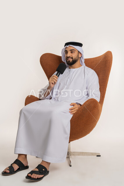 ايماءات وتعابير الاندماج والتركيز، اتقان فن الالقاء والخطابة، مهارات التحدث والحوار، بورتريه لشاب عربي خليجي اماراتي يرتدي الكندورة والغترة يجلس على الكرسي يقوم بتقديم ورش العمل والدورات التدريبية باستخدام مكبر الصوت، خلفيه بيضاء