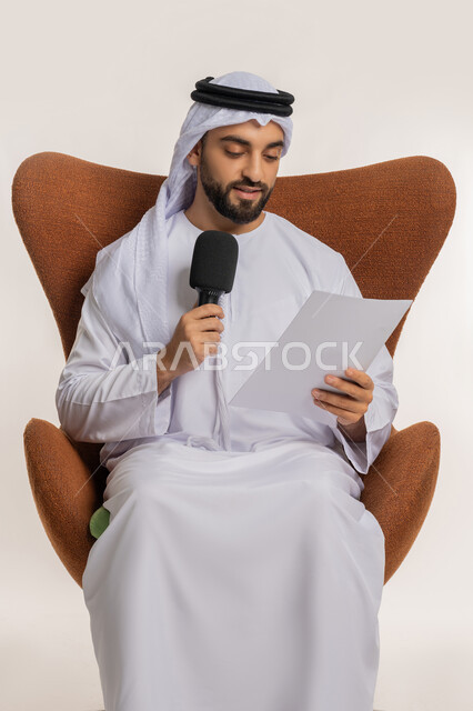 تقديم ورش العمل والدورات التدريبية، فن التحدث والخطابة، القاء الخطابات من الورقة بإيماءات الثقة بالنفس، التواصل الفعال وتبادل الحوار والنقاش، بورتريه لشاب عربي خليجي اماراتي يرتدي الكندورة والغترة يجلس على الكرسي يمسك مكبر الصوت، خلفية بيضاء